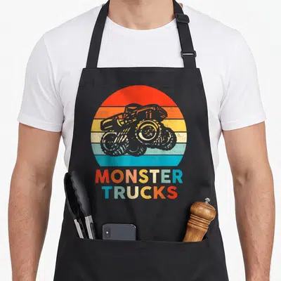 Toddler truck aprons - Grey monster truck over red orange yellow blue stripes with stacked text — shown on a black cotton canvas apron against a white background