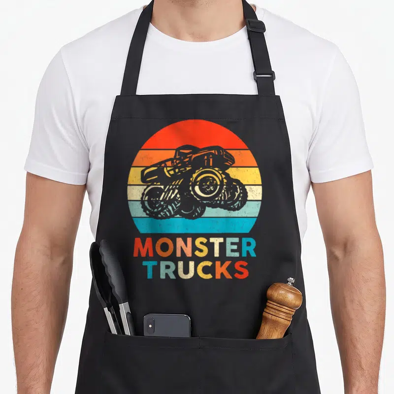 Toddler truck aprons - Grey monster truck over red orange yellow blue stripes with stacked text — shown on a black cotton canvas apron against a white background
