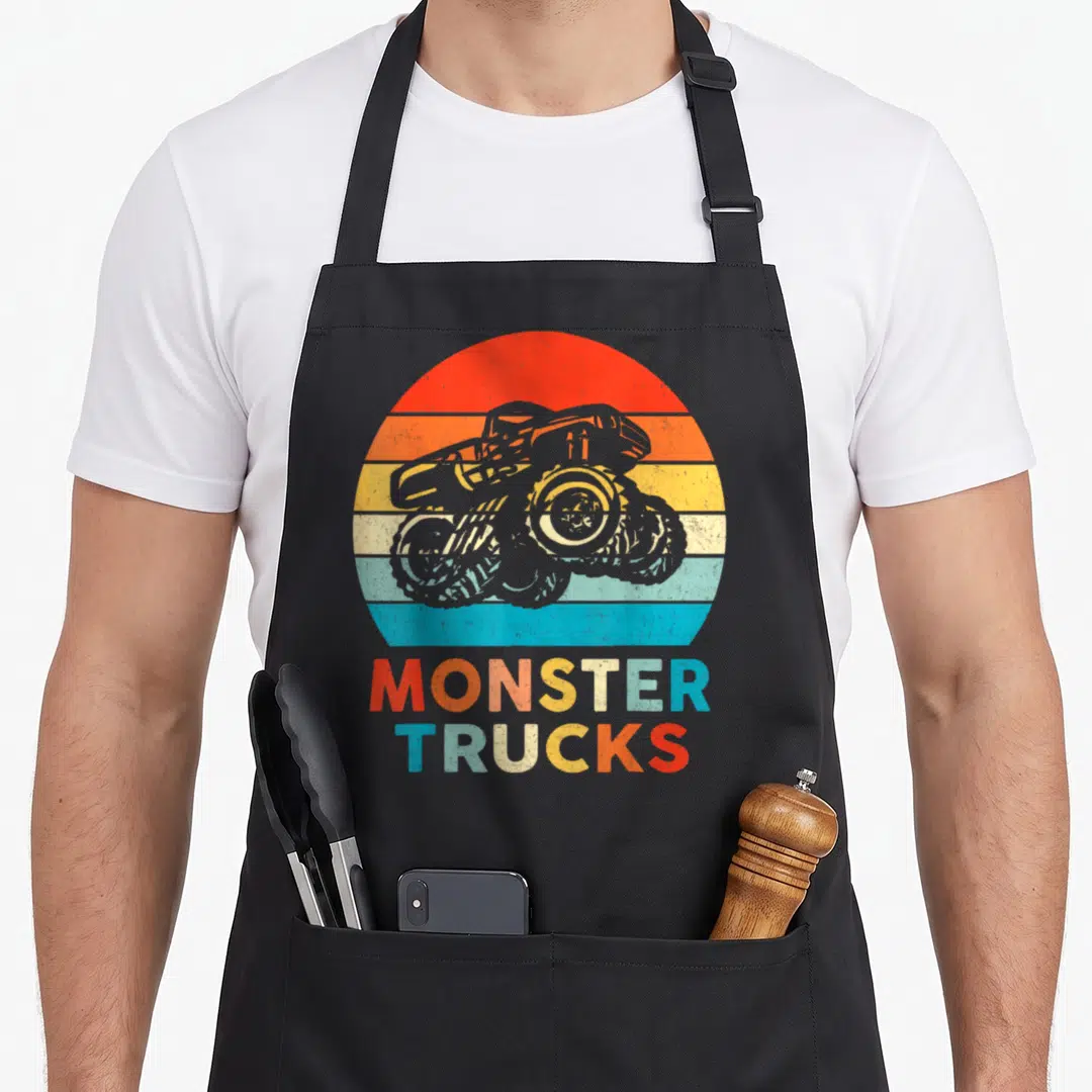 Toddler truck aprons - Grey monster truck over red orange yellow blue stripes with stacked text — shown on a black cotton canvas apron against a white background