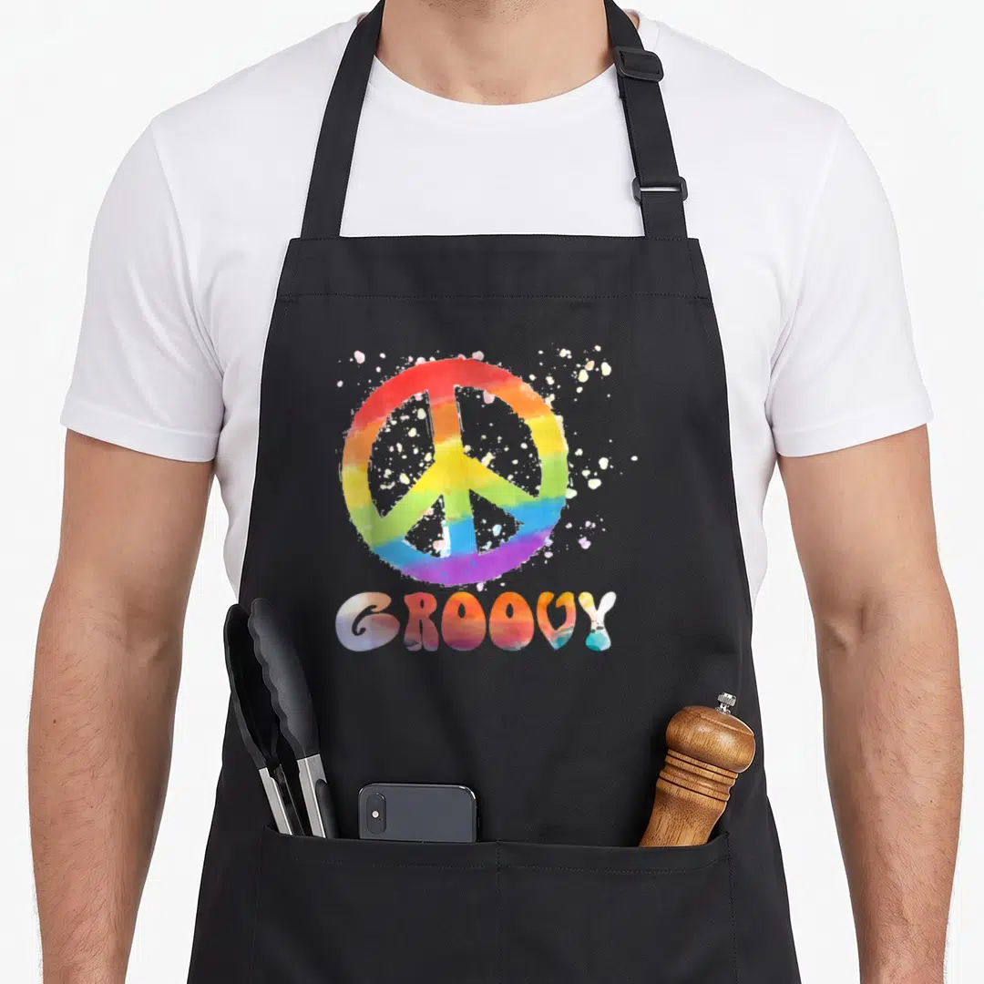 Peace 60's style aprons - Rainbow watercolor peace symbol and groovy text on cotton canvas — shown on a black cotton canvas apron against a white background