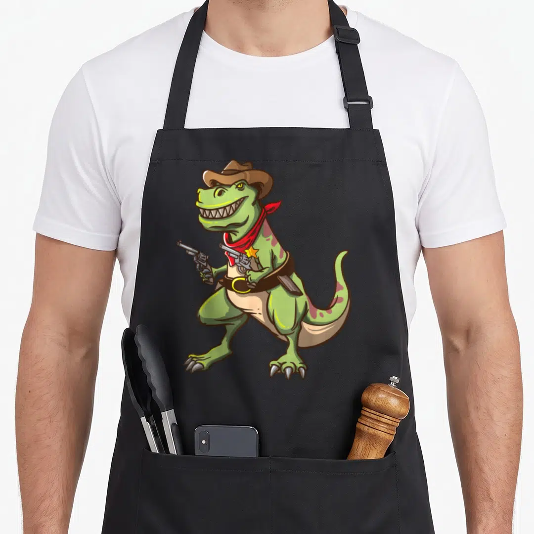 Rex cowboy apron - Cartoon green T-Rex wearing brown cowboy hat and red bandana holding two silver revolvers — shown on a black cotton canvas apron against a white background