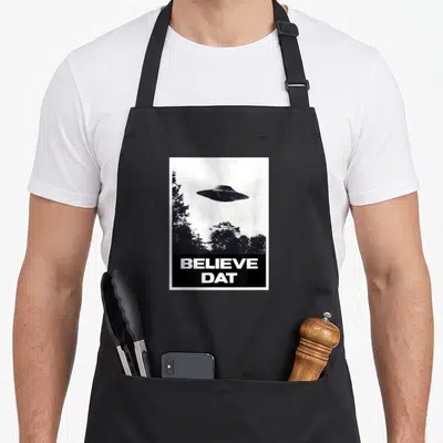 Believe ufo apron - UFO hovering over trees with BELIEVE DAT text on a black bar — shown on a black cotton canvas apron against a white background