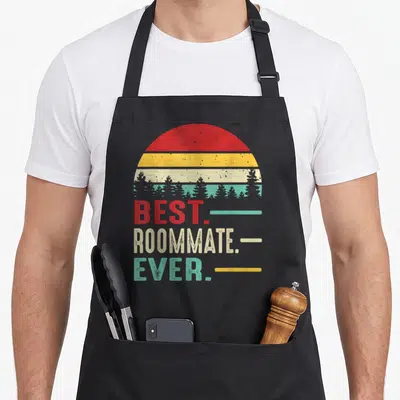 Roommate birthday apron - Retro striped sun with tree silhouettes above best roommate ever text in red yellow teal beige — shown on a black cotton canvas apron against a white background