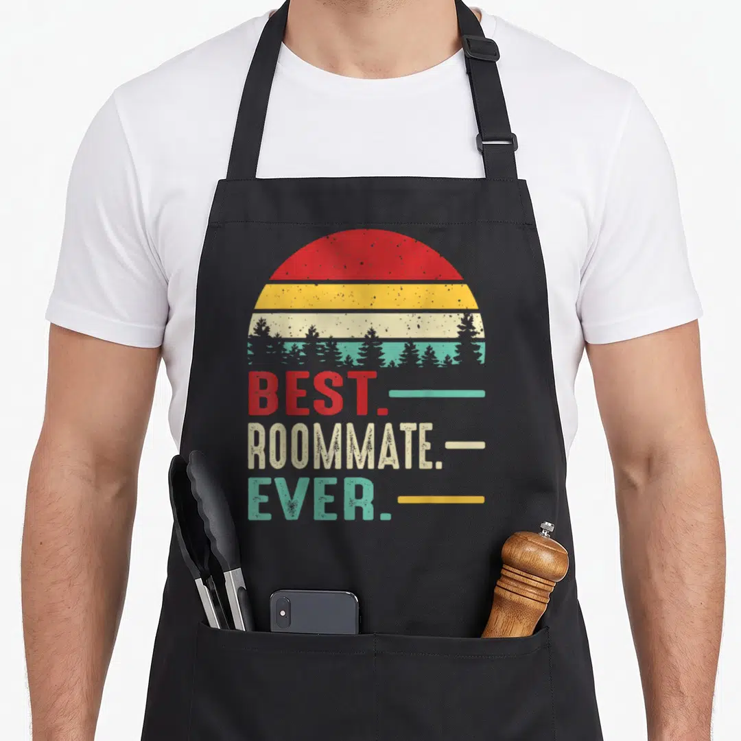 Roommate birthday apron - Retro striped sun with tree silhouettes above best roommate ever text in red yellow teal beige — shown on a black cotton canvas apron against a white background
