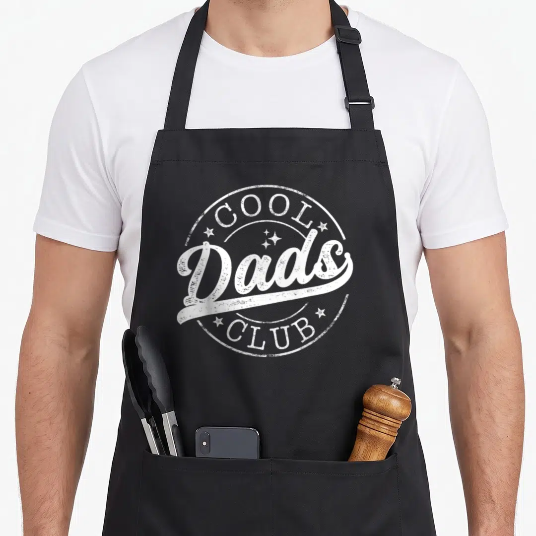 Emblem father day apron - Distressed white circular stamp reading Cool Dads Club with scattered stars — shown on a black cotton canvas apron against a white background