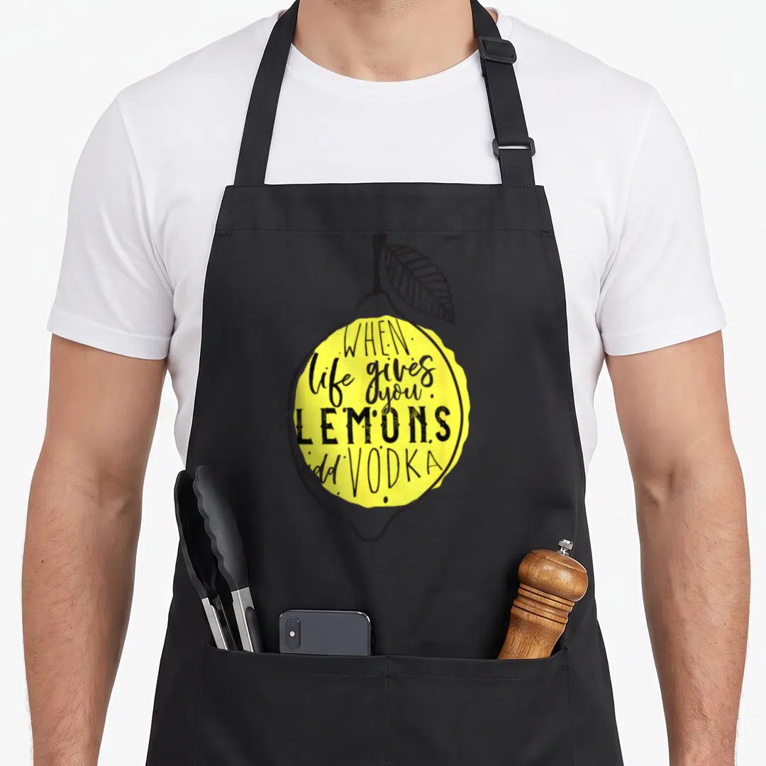 Add lemon apron - Yellow lemon with black leaf and text WHEN life gives you LEMONS add VODKA — shown on a black cotton canvas apron against a white background