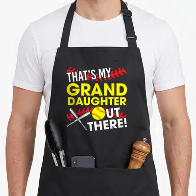 Grand grandma apron - Stacked text That's My Grand Daughter Out There with yellow softball and crossed white bats — shown on a black cotton canvas apron against a white background
