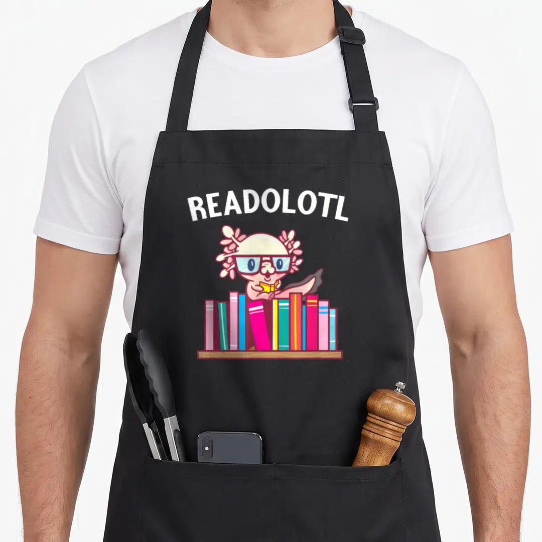 Axolotl book apron - Arched READOLOTL text over pink bespectacled axolotl holding yellow book above colorful spines — shown on a black cotton canvas apron against a white background