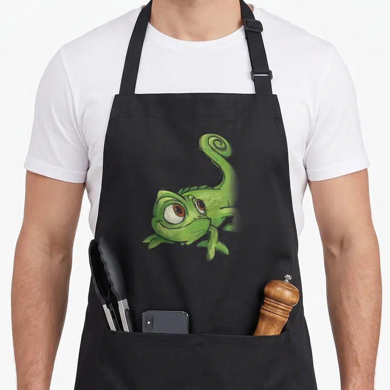 Pascal adorable aprons - Green cartoon chameleon with curled tail and large eyes centered on fabric — shown on a black cotton canvas apron against a white background