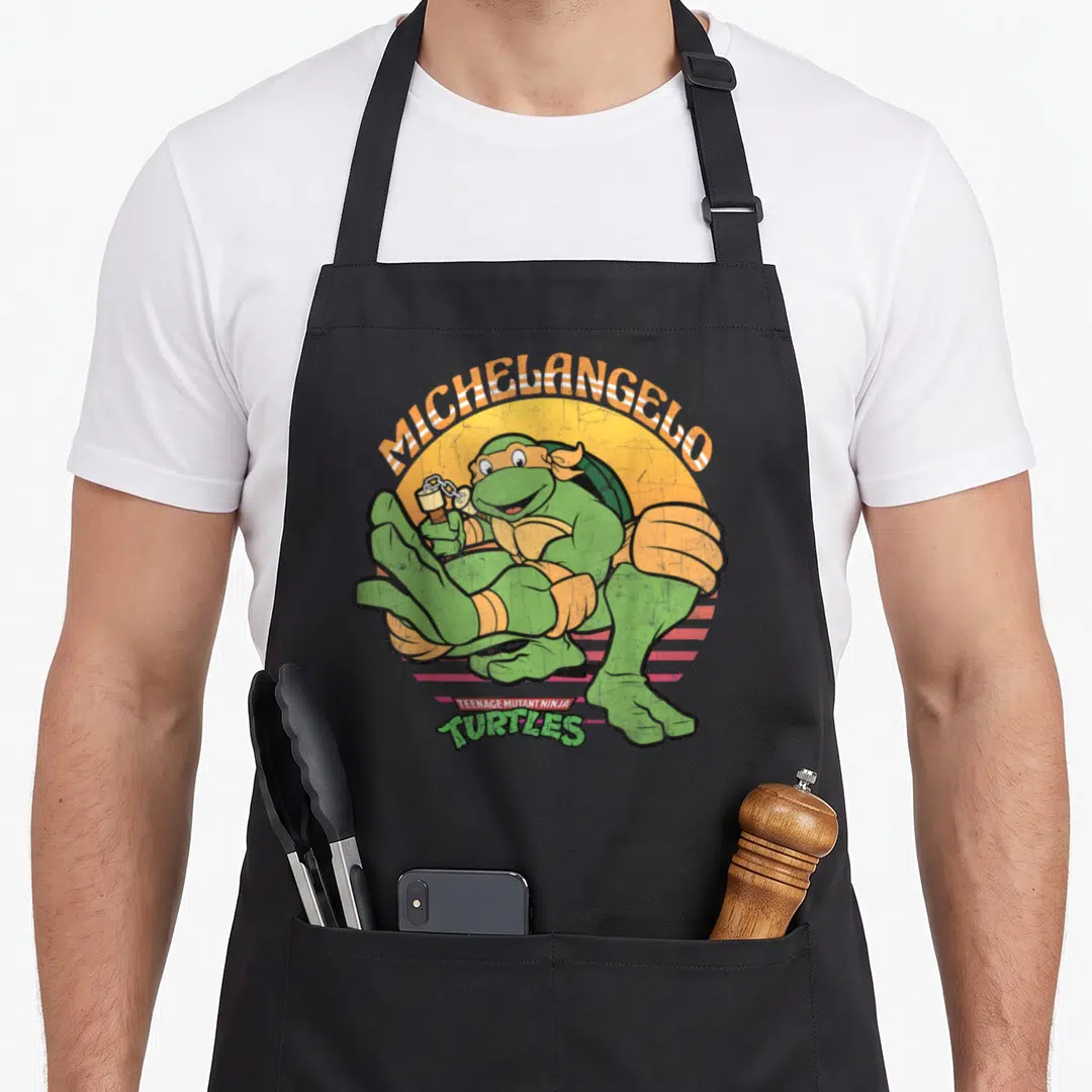 Michelangelo teenage cooking aprons - Green turtle with nunchucks, yellow sun, pink stripes, and arched orange text — shown on a black cotton canvas apron against a white background