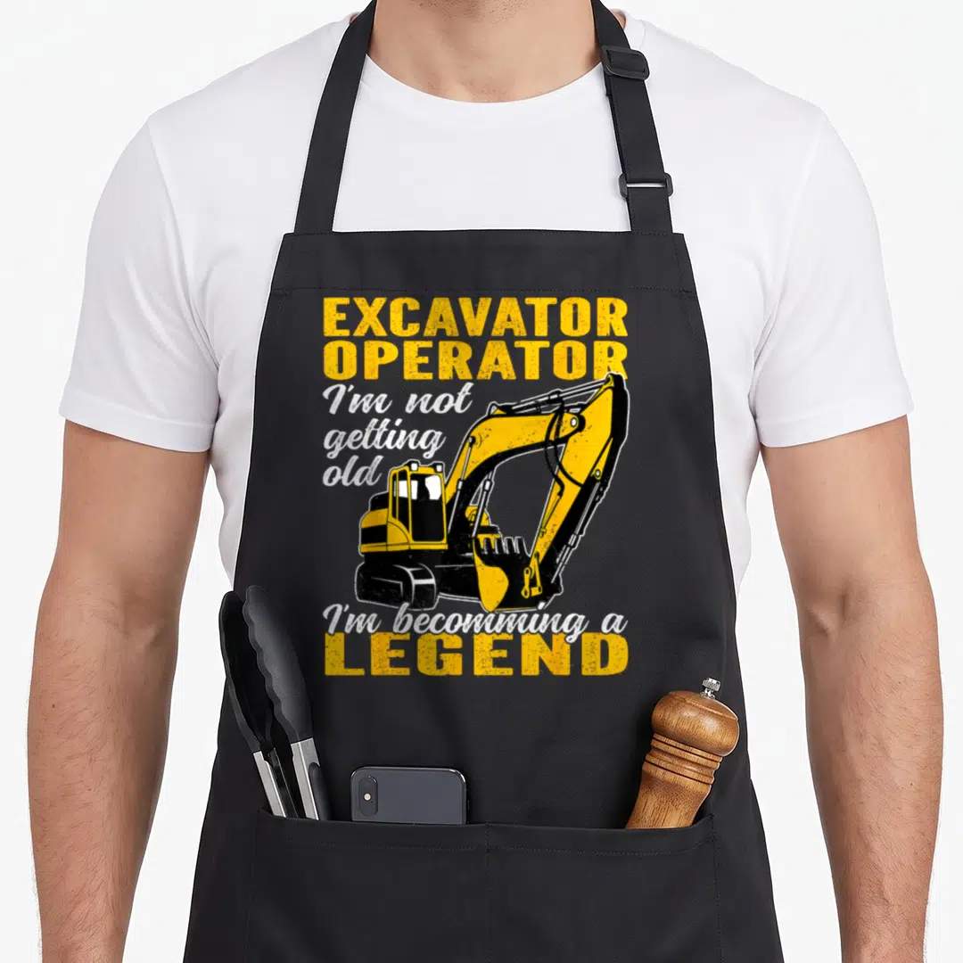 Becomming heavy canvas aprons - Yellow excavator illustration with EXCAVATOR OPERATOR and I'm becoming a LEGEND text — shown on a black cotton canvas apron against a white background