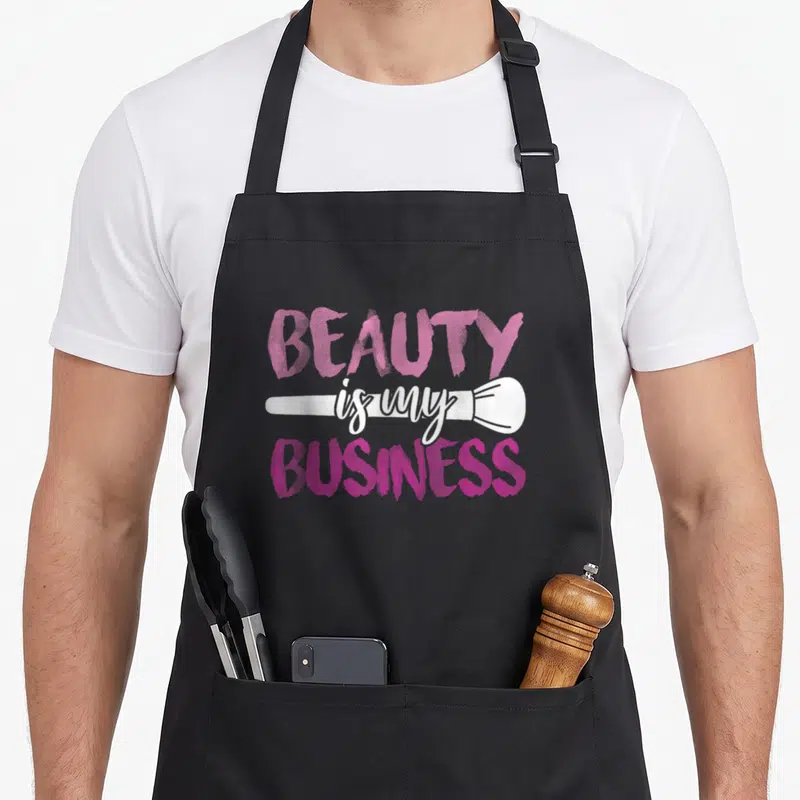 Beauty business apron - Pink and white stacked text with horizontal white makeup brush graphic — shown on a black cotton canvas apron against a white background