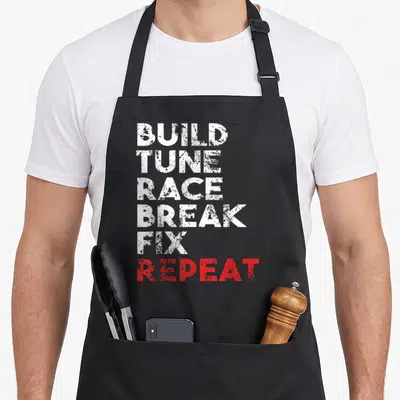 Tune car apron - Stacked white and red distressed text reading Build Tune Race Break Fix Repeat — shown on a black cotton canvas apron against a white background