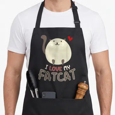 Chonk kitten cat apron - Round beige cartoon cat with red heart and I LOVE MY FATCAT text — shown on a black cotton canvas apron against a white background