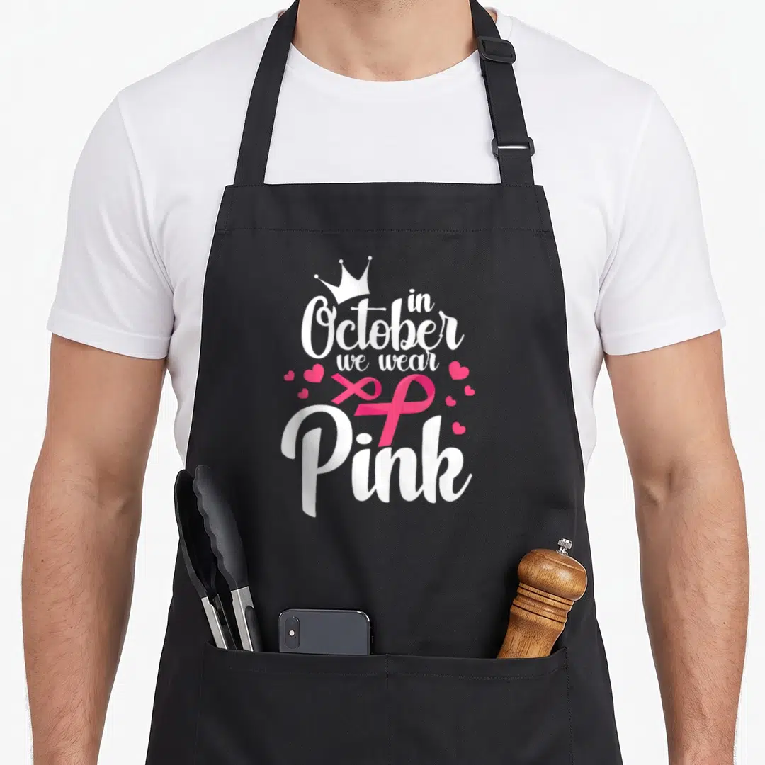 Funny october breast cancer awareness aprons - White text reading in October we wear Pink above a pink ribbon and small — shown on a black cotton canvas apron against a white background