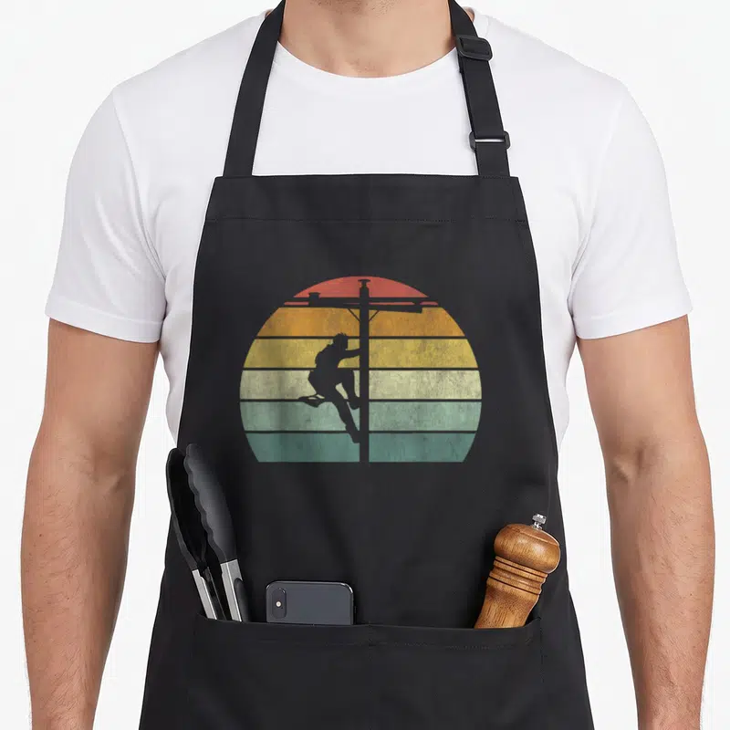 Lineman utility apron - Grey silhouette of person climbing utility pole against striped red orange yellow teal retro sun — shown on a black cotton canvas apron against a white background