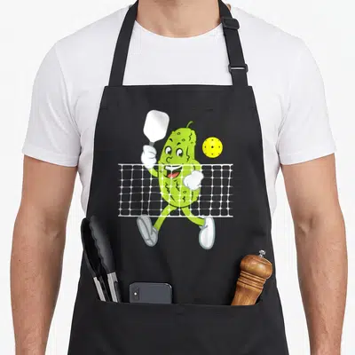 Playing pickle apron - Cartoon green pickle holding a paddle behind a white net next to a yellow perforated ball — shown on a black cotton canvas apron against a white background