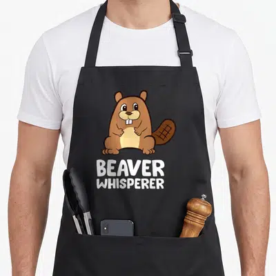 Whisperer beaver apron - Cartoon brown beaver sitting above white text BEAVER WHISPERER — shown on a black cotton canvas apron against a white background