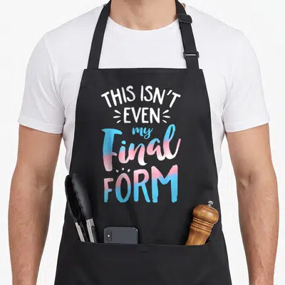 Even pride apron - Stacked text THIS ISN'T EVEN my Final FORM in trans flag blue and pink gradients — shown on a black cotton canvas apron against a white background