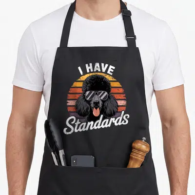 Dog breed standard size of apron - Black poodle in sunglasses with I Have Standards text over retro striped sunset — shown on a black cotton canvas apron against a white background