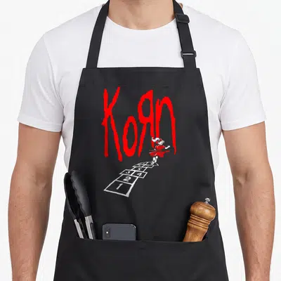Korn hop apron - Girl in red dress jumping on white hopscotch grid under large red Korn text — shown on a black cotton canvas apron against a white background