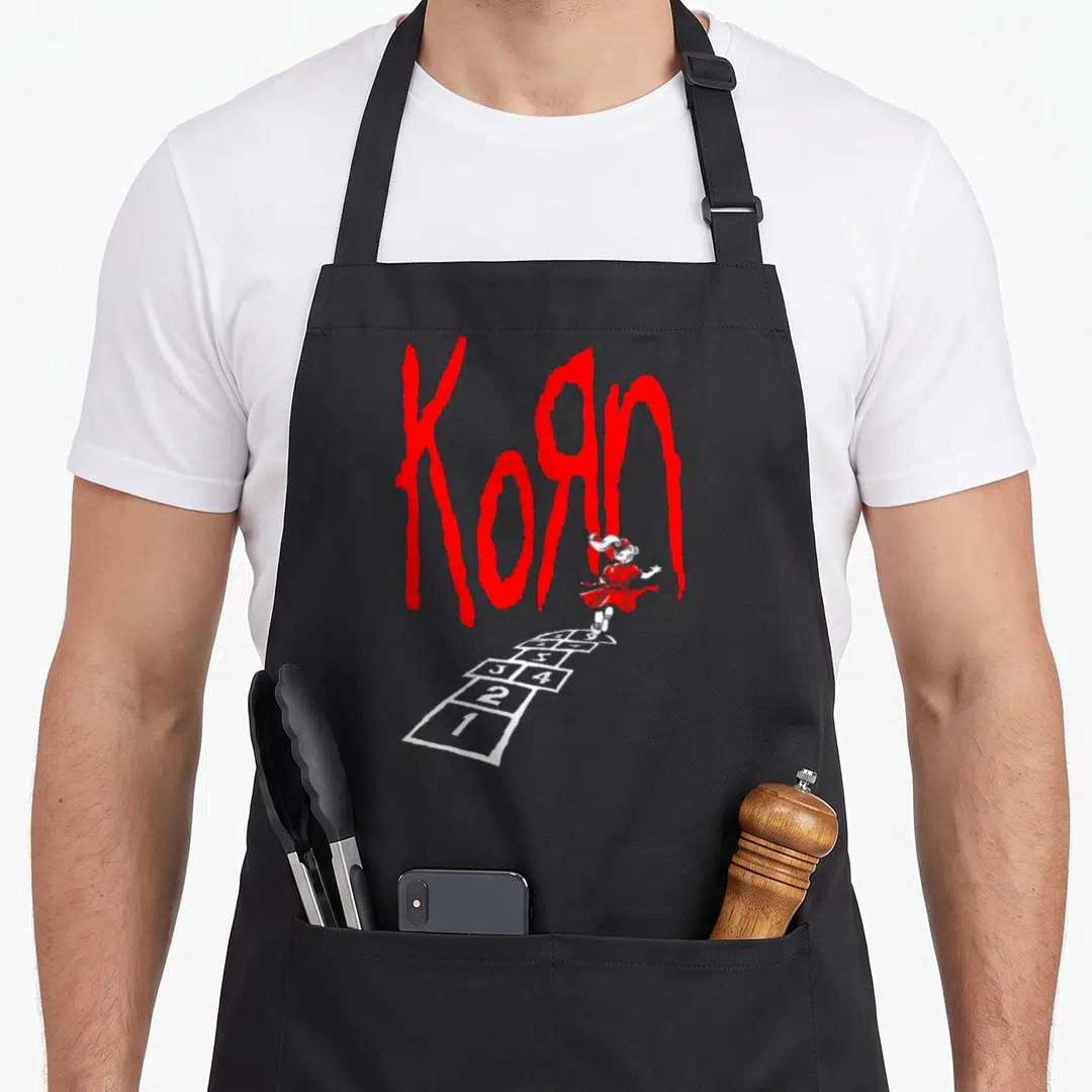 Korn hop apron - Girl in red dress jumping on white hopscotch grid under large red Korn text — shown on a black cotton canvas apron against a white background