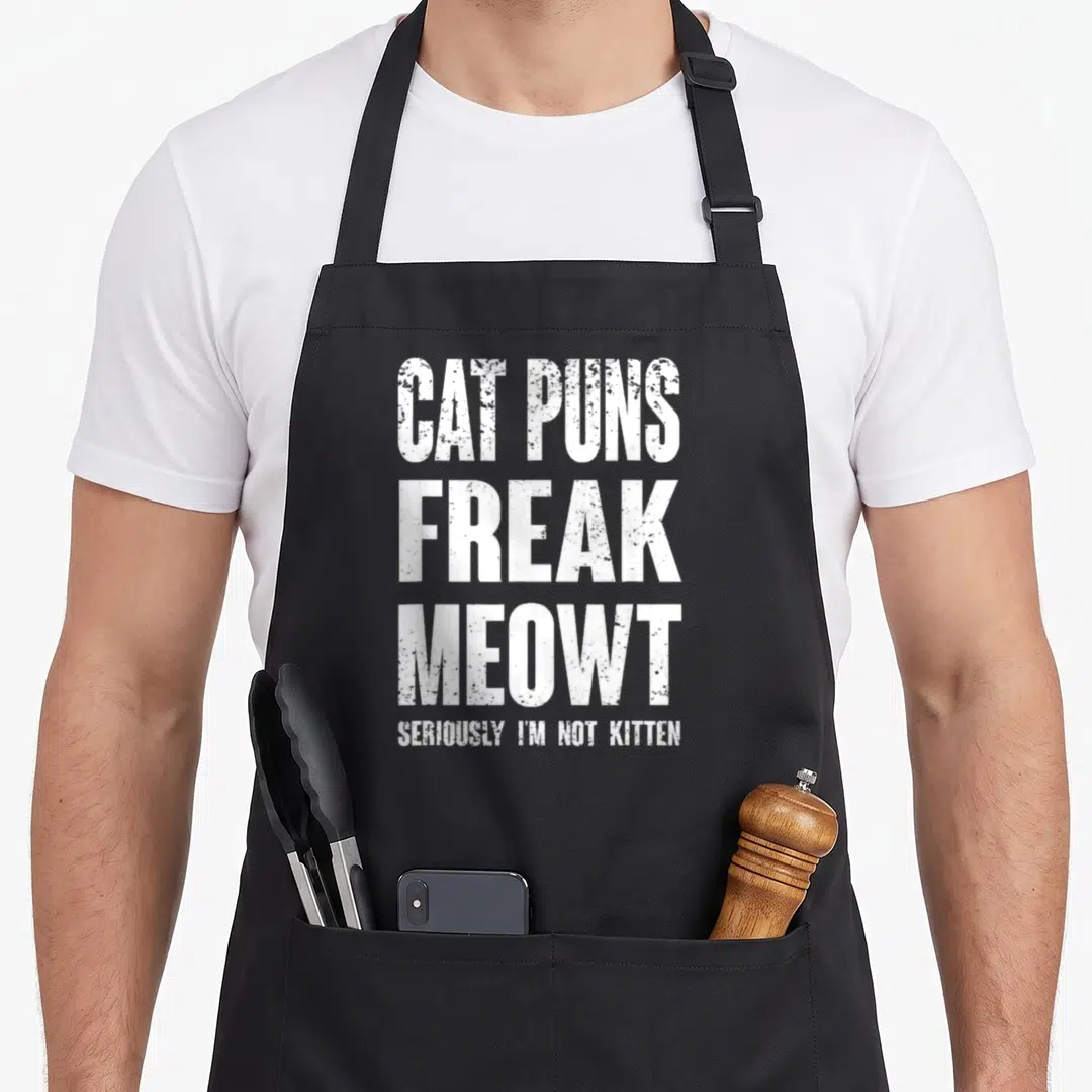Pun cat apron - White stacked text reading CAT PUNS FREAK MEOWT SERIOUSLY I'M NOT KITTEN — shown on a black cotton canvas apron against a white background