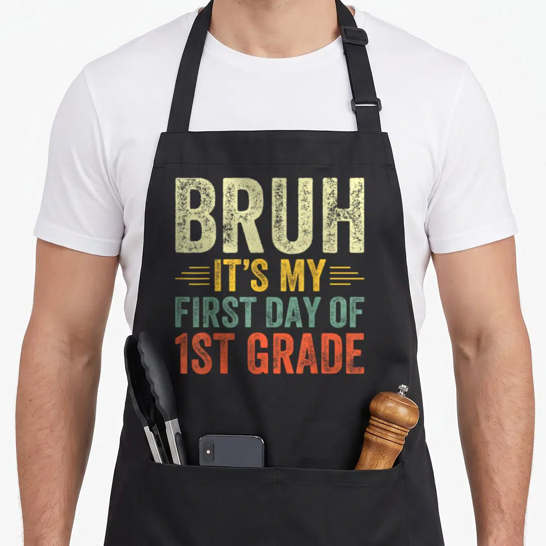 Bruh 1st teacher apron - Stacked distressed text BRUH IT'S MY FIRST DAY OF 1ST GRADE in beige yellow green orange — shown on a black cotton canvas apron against a white background