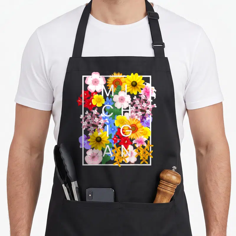 Michigander pride apron - Vertical white MICHIGAN text surrounded by yellow, red, pink, blue, and green flowers — shown on a black cotton canvas apron against a white background