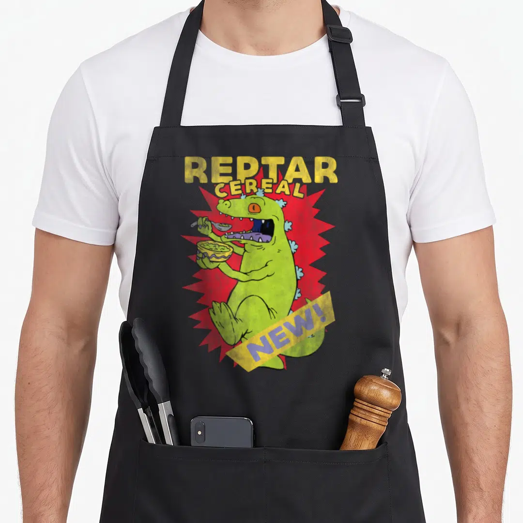 Reptar cereal apron - Green dinosaur eating cereal from yellow bowl over red burst with REPTAR CEREAL NEW! text — shown on a black cotton canvas apron against a white background