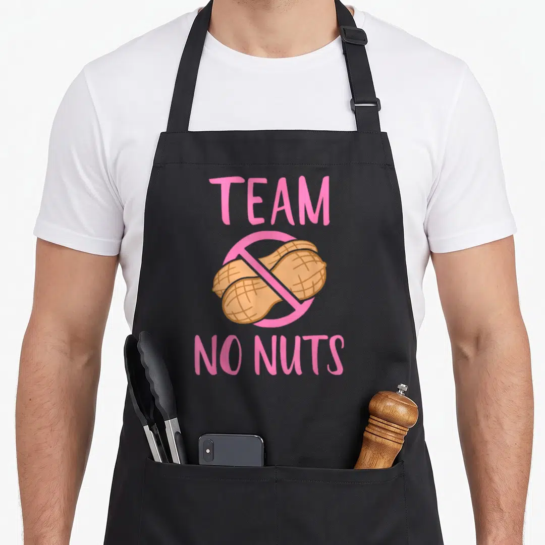 Nut crew apron - Pink TEAM NO NUTS text with two tan peanuts crossed out in prohibition sign — shown on a black cotton canvas apron against a white background