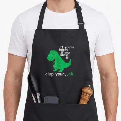Clap t-rex apron - Green cartoon dinosaur with text If you're happy & you know it clap your oh — shown on a black cotton canvas apron against a white background
