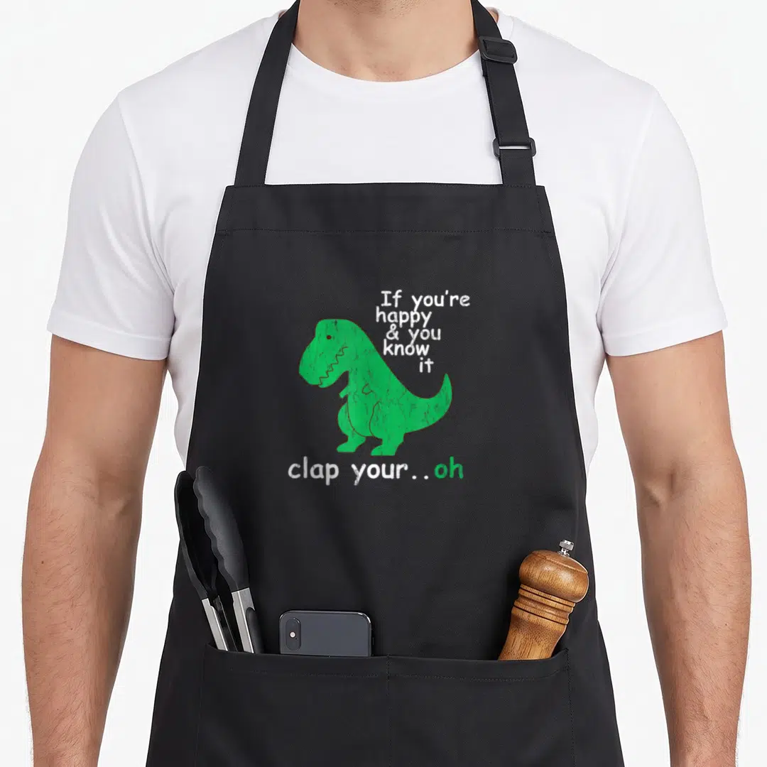 Clap t-rex apron - Green cartoon dinosaur with text If you're happy & you know it clap your oh — shown on a black cotton canvas apron against a white background