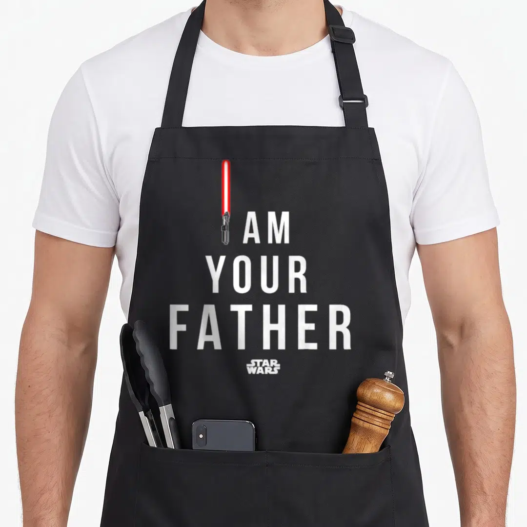 Funny father falcon dad apron - I AM YOUR FATHER text with red lightsaber replacing the I and Star Wars logo below — shown on a black cotton canvas apron against a white background