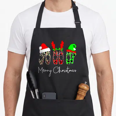 Ballet merry christmas apron - Three patterned ballet shoes with Santa hat, reindeer antlers, and elf hat above Merry — shown on a black cotton canvas apron against a white background