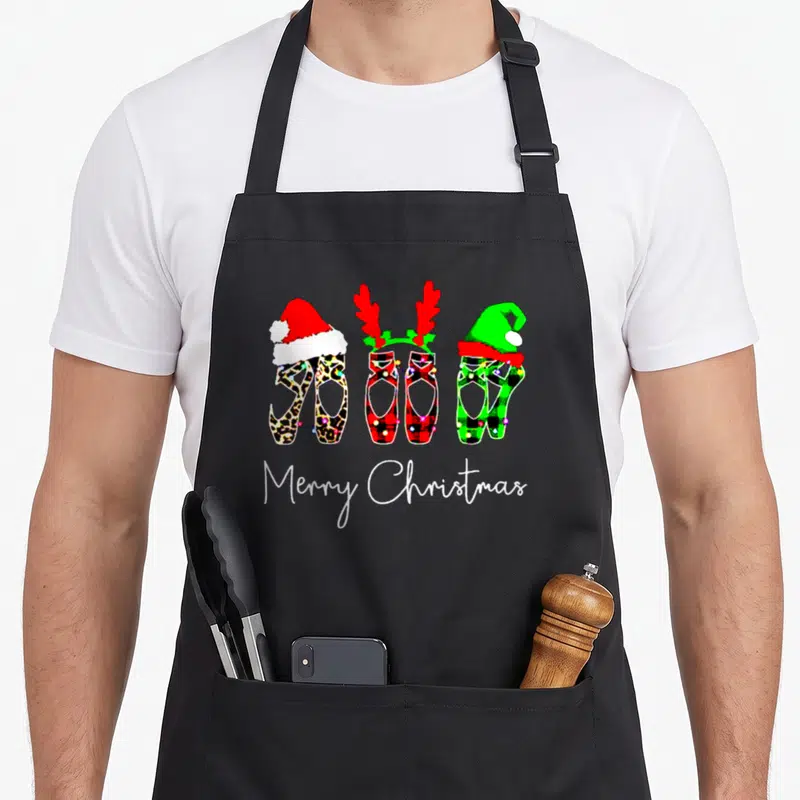 Ballet merry christmas apron - Three patterned ballet shoes with Santa hat, reindeer antlers, and elf hat above Merry — shown on a black cotton canvas apron against a white background