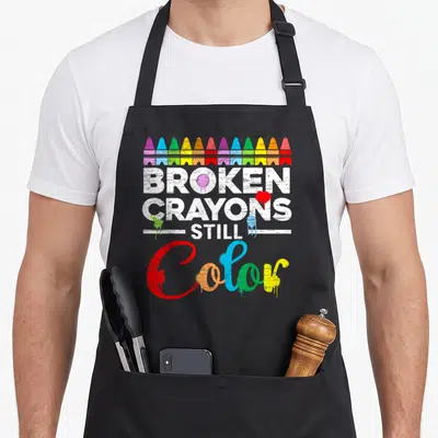Mental crayons apron - Row of broken crayons above distressed white text and multi-colored dripping script reading Color — shown on a black cotton canvas apron against a white background