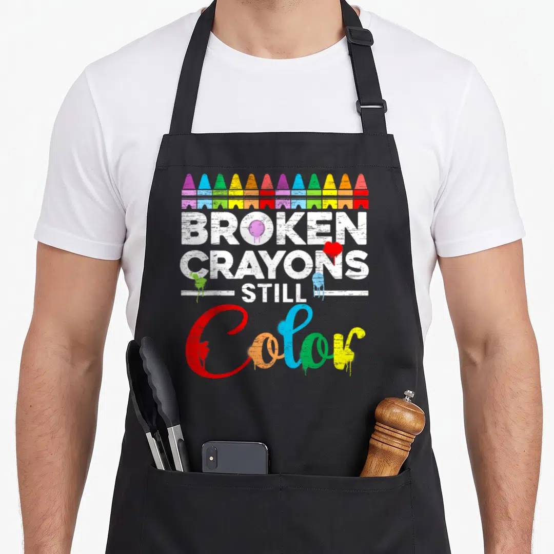 Mental crayons apron - Row of broken crayons above distressed white text and multi-colored dripping script reading Color — shown on a black cotton canvas apron against a white background