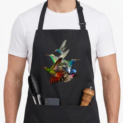 Hummingbird bird apron - Five realistic hummingbirds in blue, green, red, and brown clustered in flight — shown on a black cotton canvas apron against a white background