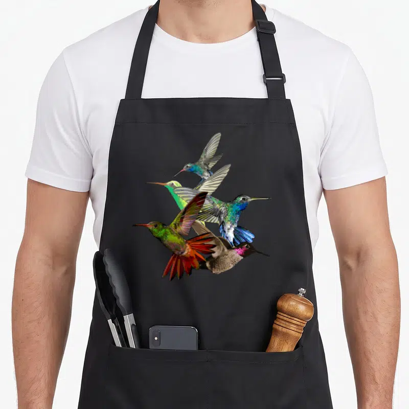 Hummingbird bird apron - Five realistic hummingbirds in blue, green, red, and brown clustered in flight — shown on a black cotton canvas apron against a white background