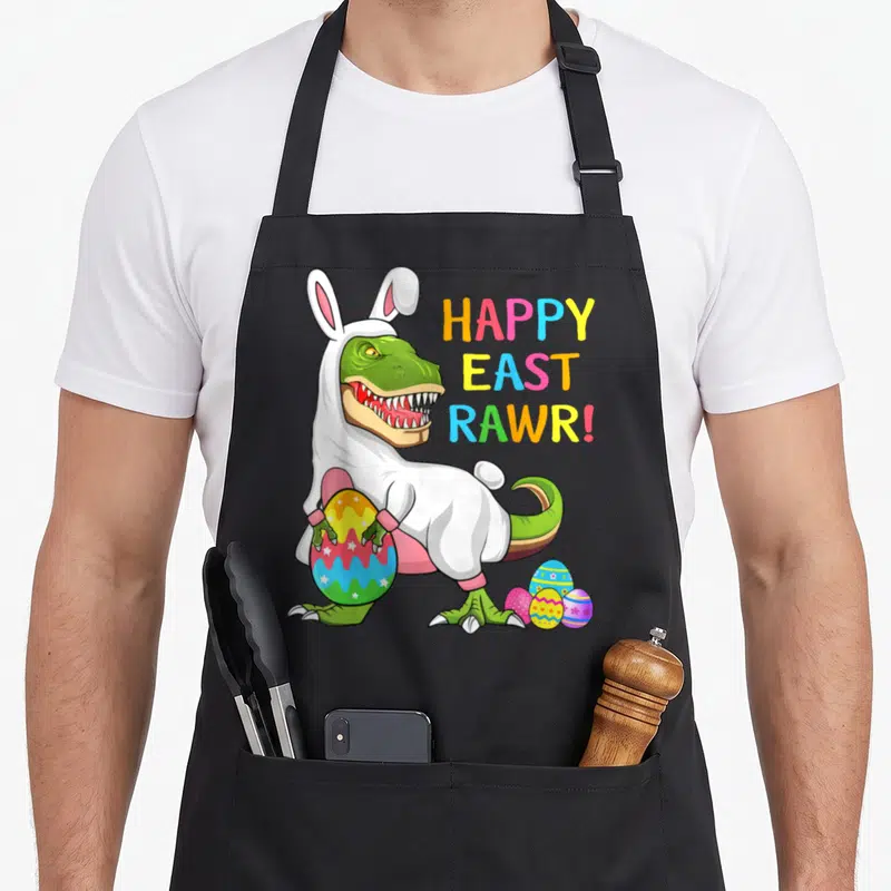 Dinosaur easter apron - Green T-Rex in white bunny suit holding large egg with HAPPY EAST RAWR! text — shown on a black cotton canvas apron against a white background