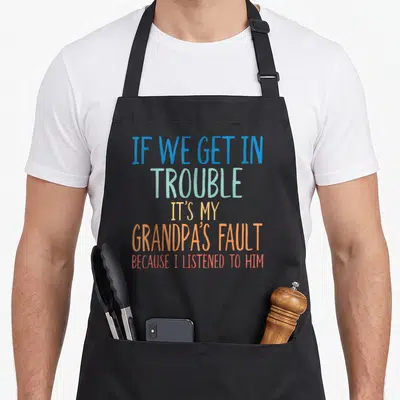 Trouble grandson apron - Stacked colorful text reading IF WE GET IN TROUBLE IT'S MY GRANDPA'S FAULT BECAUSE I LISTENED — shown on a black cotton canvas apron against a white background