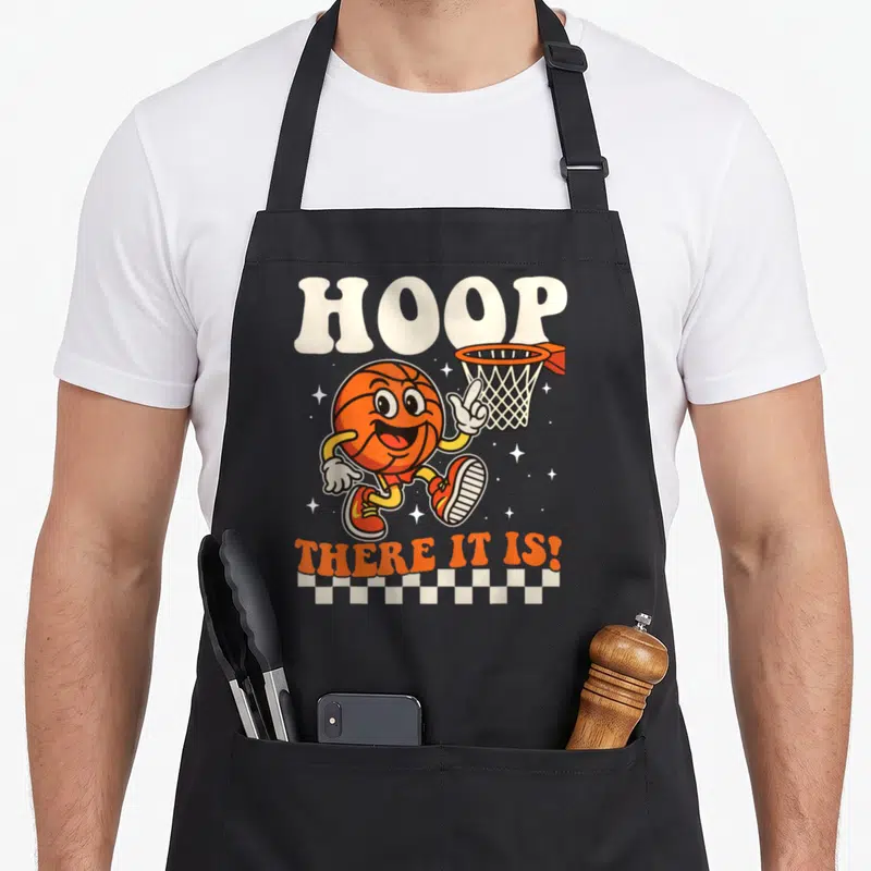 Hoop basketball back apron - Cartoon orange basketball pointing to hoop with HOOP THERE IT IS! text and checkered border — shown on a black cotton canvas apron against a white background