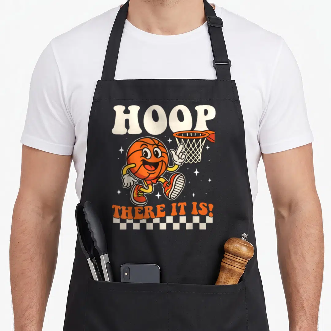 Hoop basketball back apron - Cartoon orange basketball pointing to hoop with HOOP THERE IT IS! text and checkered border — shown on a black cotton canvas apron against a white background