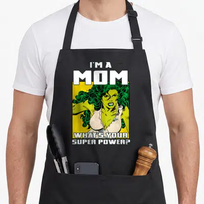 She mother's day aprons - Cartoon green woman yellow background I'M A MOM WHAT'S YOUR SUPER POWER text — shown on a black cotton canvas apron against a white background