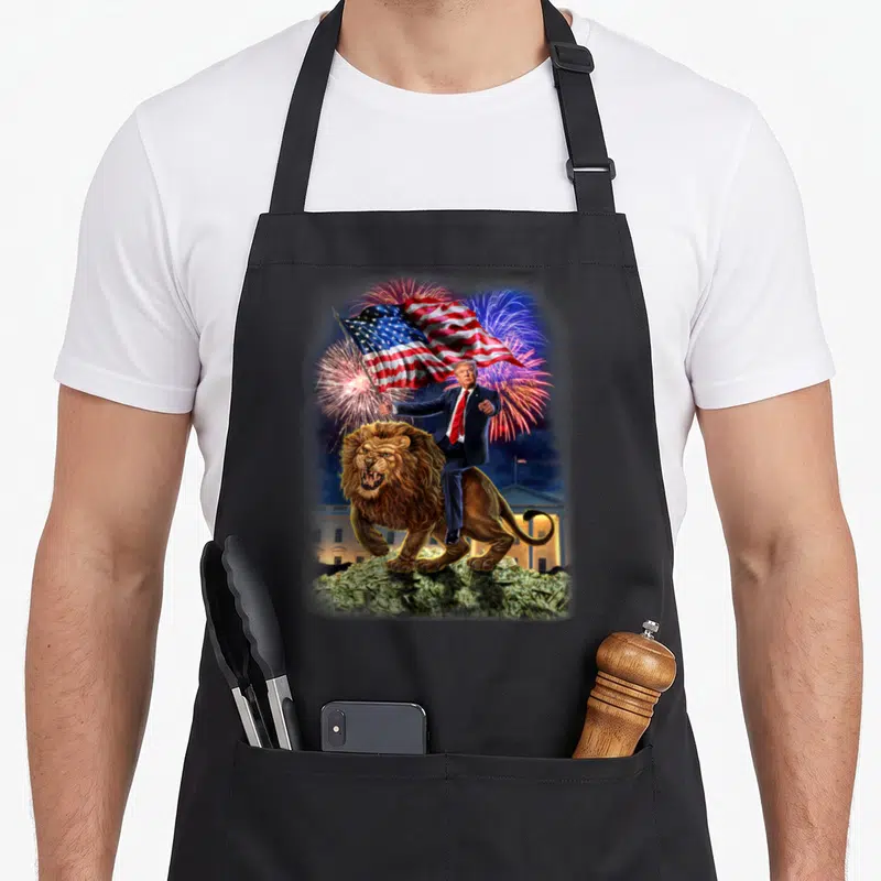 Republican lion apron - Trump riding lion holding USA flag over money pile with fireworks — shown on a black cotton canvas apron against a white background