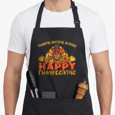 Grateful thankful apron - Cartoon turkey in hat surrounded by pumpkins and autumn leaves with holiday text — shown on a black cotton canvas apron against a white background