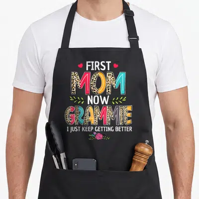 Grammie mother's day aprons - Stacked text First Mom Now Grammie with hearts leaves and floral print on black apron — shown on a black cotton canvas apron against a white background