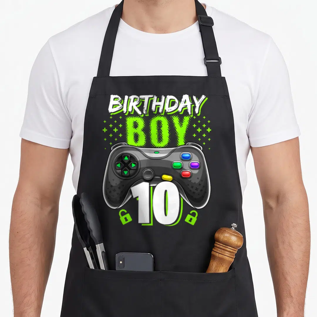 Birthday controller gamer video game apron - Cartoon game controller with BIRTHDAY BOY 10 text and scattered plus signs — shown on a black cotton canvas apron against a white background