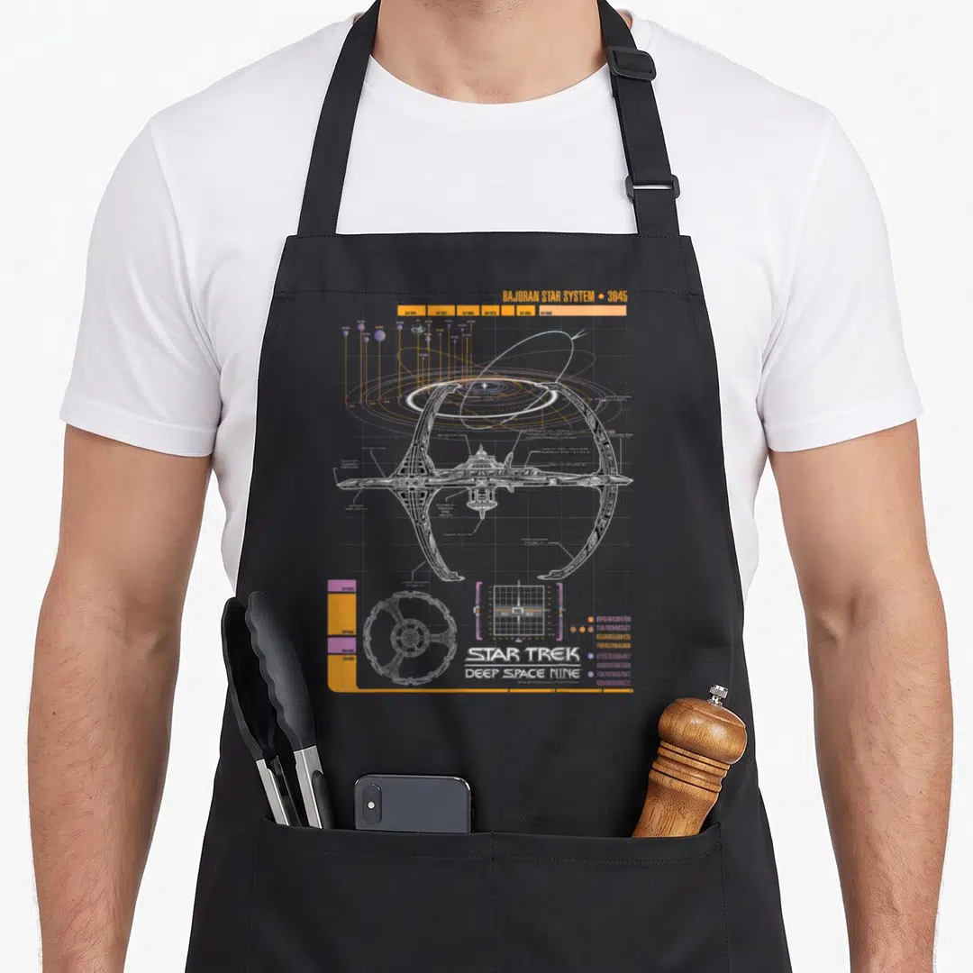 Ds9 star apron - Grey and orange technical schematic of a space station on a grid with orbital paths — shown on a black cotton canvas apron against a white background